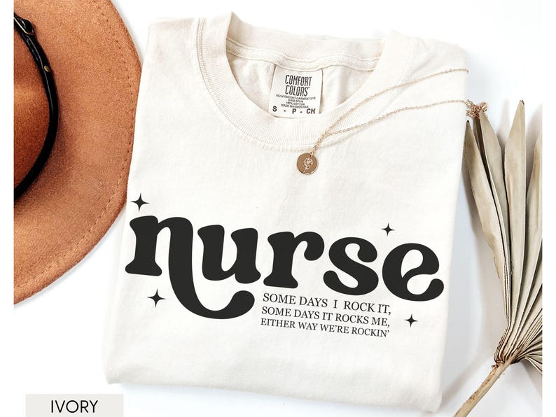 Pode incluir: Camiseta cor marfim com a palavra "nurse" em negrito preto, acompanhada do texto "Some days I rock it, some days it rocks me, either way we're rockin'". Um colar dourado e um chap&eacute;u marrom tamb&eacute;m s&atilde;o vis&iacute;veis.