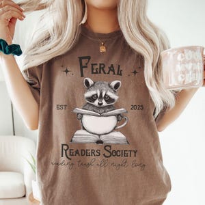 May include: A brown t-shirt with a graphic of a raccoon reading a book and drinking coffee. The text on the shirt reads "Feral Readers Society est. 2025 reading trash all night long".