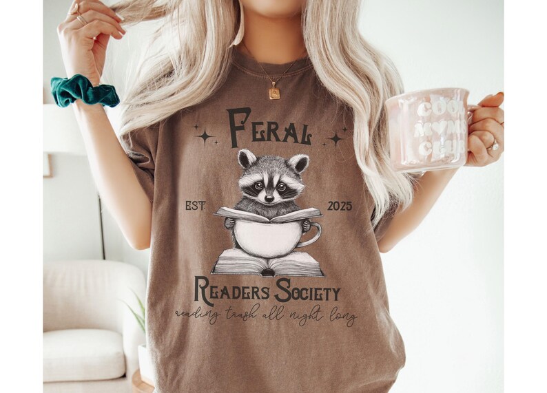 May include: A brown t-shirt with a graphic of a raccoon reading a book and drinking coffee. The text on the shirt reads "Feral Readers Society est. 2025 reading trash all night long".