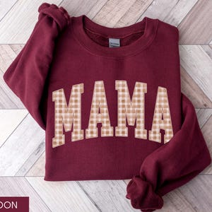 May include: Maroon sweatshirt with the word "MAMA" in large, tan and white gingham letters across the front. The sweatshirt is folded on a light wood-grain surface. The sleeves are folded up.