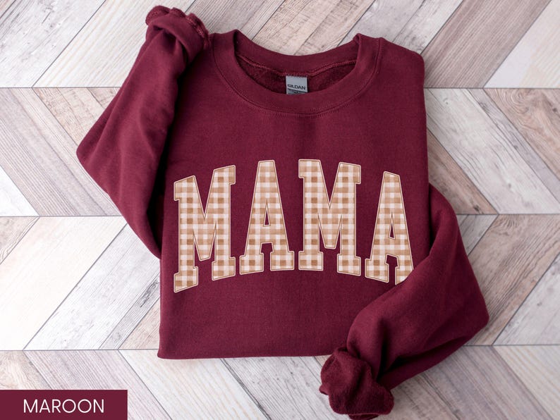 May include: Maroon sweatshirt with the word "MAMA" in large, tan and white gingham letters across the front. The sweatshirt is folded on a light wood-grain surface. The sleeves are folded up.