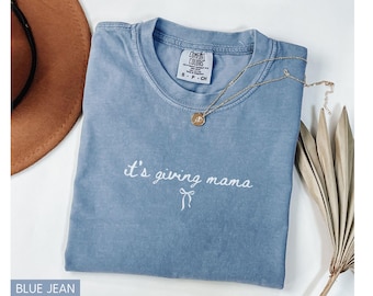 May include: A blue jean colored t-shirt with the phrase "it's giving mama" embroidered in white. A gold necklace with a pendant is draped across the shirt. A brown hat and dried plant stems are in the background.