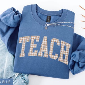 May include: Indigo blue sweatshirt with the word "TEACH" in a tan and white plaid design. A silver necklace with a small pendant is draped across the sweatshirt. The sweatshirt is folded on top of a pair of blue jeans.