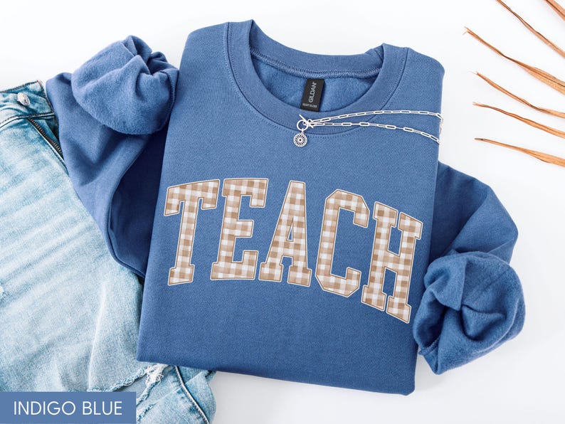 May include: Indigo blue sweatshirt with the word "TEACH" in a tan and white plaid design. A silver necklace with a small pendant is draped across the sweatshirt. The sweatshirt is folded on top of a pair of blue jeans.