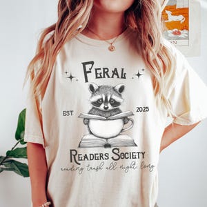 May include: A white t-shirt with a black and white illustration of a raccoon reading a book. The text on the shirt reads "Feral Readers Society est. 2025 reading trash all night long."