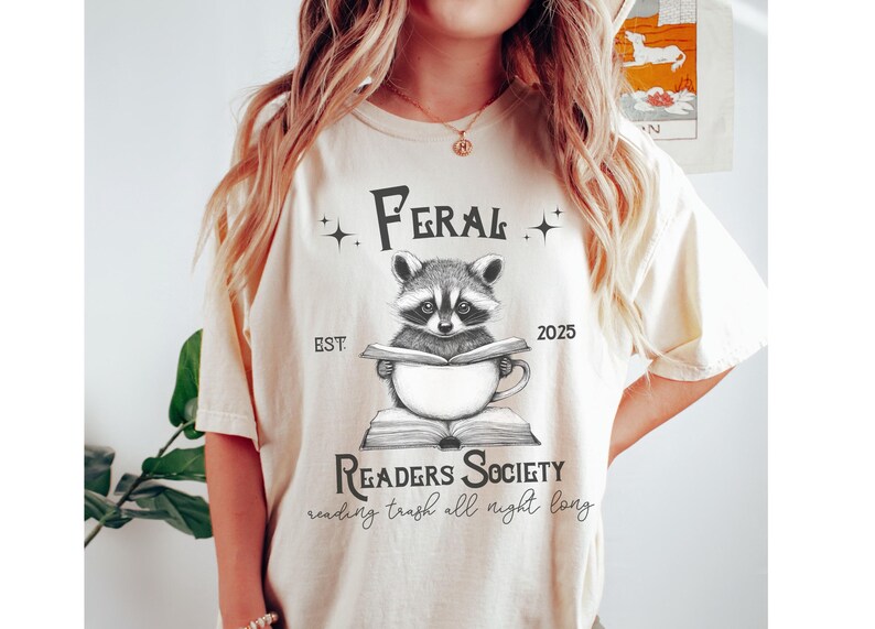 May include: A white t-shirt with a black and white illustration of a raccoon reading a book. The text on the shirt reads "Feral Readers Society est. 2025 reading trash all night long."