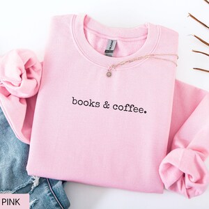 May include: A light pink sweatshirt with the text "books & coffee" in black. The sweatshirt is folded with the sleeves cuffed. A gold necklace with a small pendant rests on the sweatshirt. Blue jeans are visible in the background.