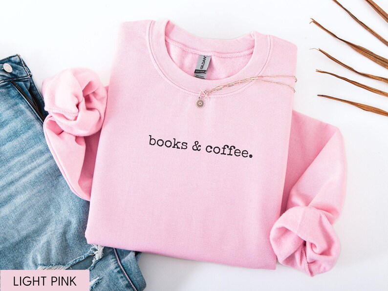 May include: A light pink sweatshirt with the text "books & coffee" in black. The sweatshirt is folded with the sleeves cuffed. A gold necklace with a small pendant rests on the sweatshirt. Blue jeans are visible in the background.