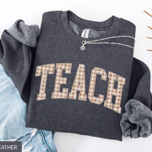 May include: Dark heather gray sweatshirt with the word "TEACH" in a tan and white plaid design. The sweatshirt is folded with the sleeves showing a gray lining. A silver necklace is draped across the top. Blue jeans are visible in the background.