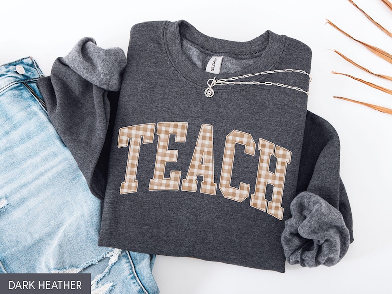 May include: Dark heather gray sweatshirt with the word "TEACH" in a tan and white plaid design. The sweatshirt is folded with the sleeves showing a gray lining. A silver necklace is draped across the top. Blue jeans are visible in the background.