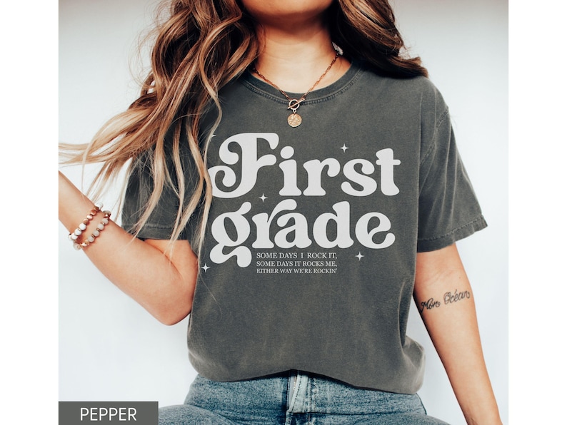 Puede incluir: Una camiseta gris oscuro con las palabras "First Grade" en blanco, con una fuente retro. La camiseta tambi&eacute;n presenta el texto "SOME DAYS I ROCK IT, SOME DAYS IT ROCKS ME, EITHER WAY WE'RE ROCKIN'" en una fuente m&aacute;s peque&ntilde;a. La camiseta es de color pimienta.