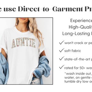 May include: A white t-shirt with the word "AUNTIE" printed in colorful letters. The text is made up of small floral patterns. The shirt is being modeled by a person wearing a denim jacket. The text "We use Direct-to-Garment Print" is at the top of the image. The text "Experience High-Quality, Long-Lasting Prints" is on the right side of the image. Below this text is a list of features of the print: "won't crack or peel", "soft fabric", "state-of-the-art print", "rated for 50+ washings". There are instructions for washing the shirt: "*wash inside out, in cold water, on gentle cycle, tumble dry low or air dry."