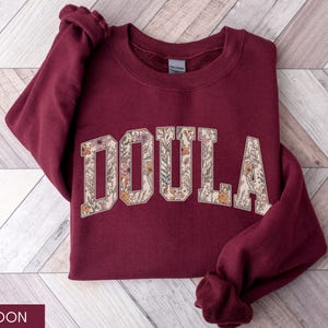 May include: Maroon sweatshirt with the word "DOULA" in a floral design. The letters are filled with a cream-colored background and various flowers in shades of pink, orange, and green. The sweatshirt is displayed on a wooden surface.