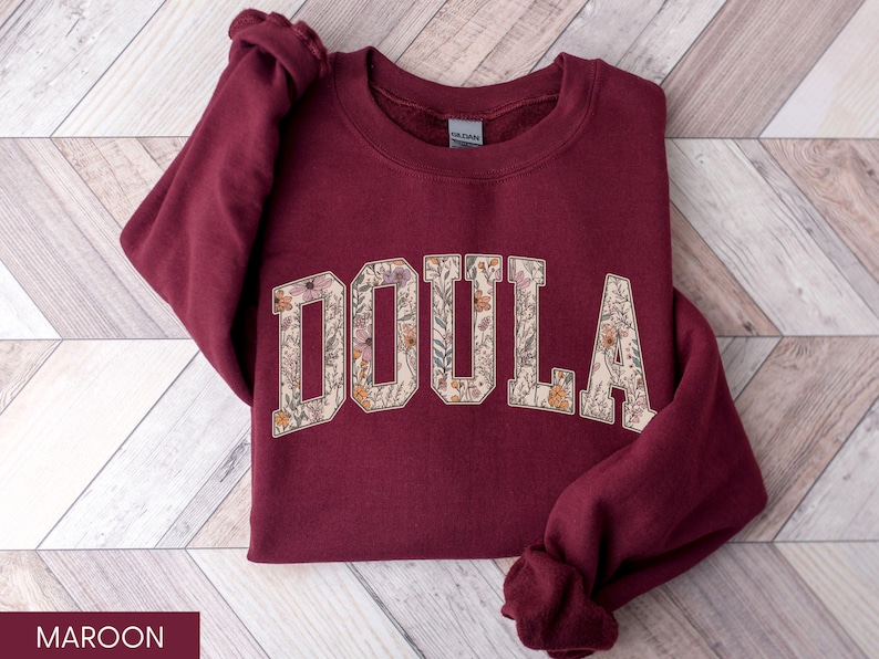 May include: Maroon sweatshirt with the word "DOULA" in a floral design. The letters are filled with a cream-colored background and various flowers in shades of pink, orange, and green. The sweatshirt is displayed on a wooden surface.