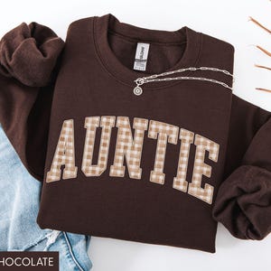 May include: Dark chocolate brown sweatshirt with the word "AUNTIE" in a plaid design. A silver necklace with a pendant is draped across the sweatshirt. The sweatshirt is paired with blue jeans.