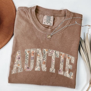 May include: A brown t-shirt with the word "AUNTIE" printed in white letters outlined with a floral pattern. The shirt is made of a soft, comfortable material and has a relaxed fit.