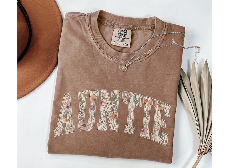 May include: A brown t-shirt with the word "AUNTIE" printed in white letters outlined with a floral pattern. The shirt is made of a soft, comfortable material and has a relaxed fit.
