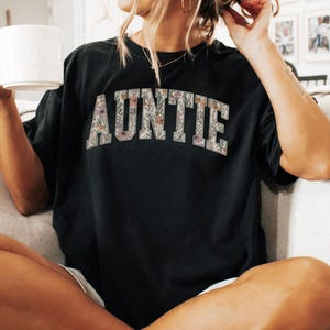 May include: A black t-shirt with the word "AUNTIE" printed in white letters with a floral pattern. The letters are outlined in black.