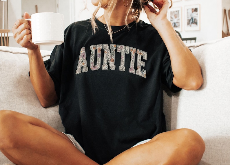 May include: A black t-shirt with the word "AUNTIE" printed in white letters with a floral pattern. The letters are outlined in black.