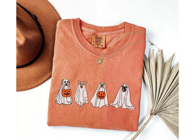 Puede incluir: Camiseta de color coral con cuatro perros disfrazados de fantasmas, cada uno sosteniendo una calabaza de Halloween. Un collar dorado y un sombrero marr&oacute;n son visibles en el fondo. La camiseta tiene una etiqueta Comfort Colors.