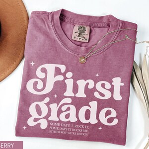 Puede incluir: Camiseta de color bayas con las palabras "First Grade" en una fuente retro. La camiseta tambi&eacute;n tiene el texto "SOME DAYS I ROCK IT, SOME DAYS IT ROCKS ME, EITHER WAY WE'RE ROCKIN". Tambi&eacute;n se ven un collar dorado y un sombrero marr&oacute;n.