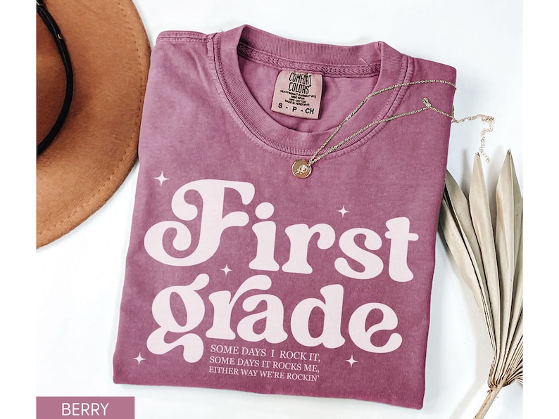 Puede incluir: Camiseta de color bayas con las palabras "First Grade" en una fuente retro. La camiseta tambi&eacute;n tiene el texto "SOME DAYS I ROCK IT, SOME DAYS IT ROCKS ME, EITHER WAY WE'RE ROCKIN". Tambi&eacute;n se ven un collar dorado y un sombrero marr&oacute;n.