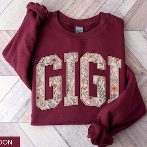 May include: Maroon sweatshirt featuring the word "GIGI" in large, floral-filled letters. The letters are outlined in cream, with orange, pink, and green flowers inside. The sweatshirt is folded on a light wood-grain surface.