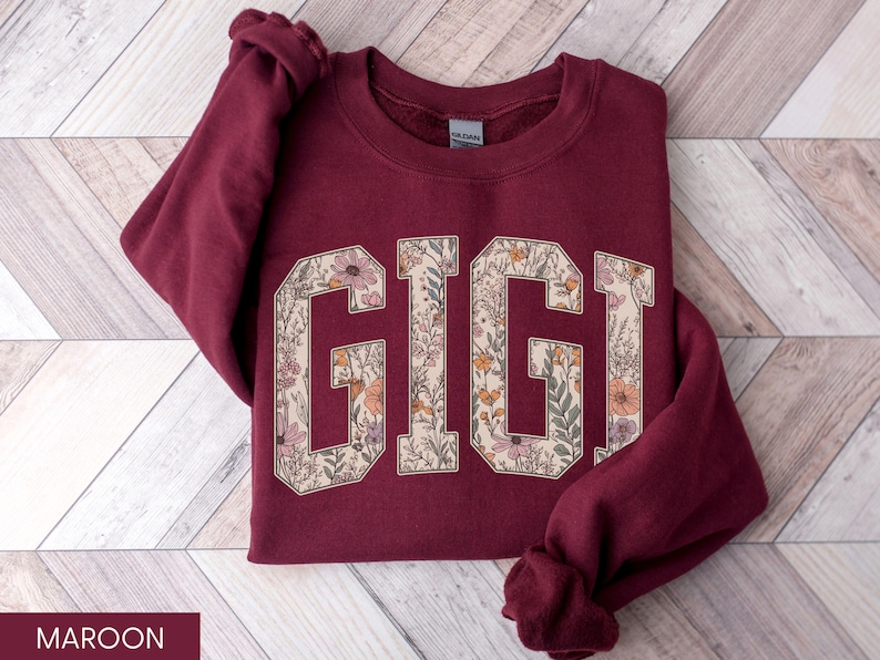 May include: Maroon sweatshirt featuring the word "GIGI" in large, floral-filled letters. The letters are outlined in cream, with orange, pink, and green flowers inside. The sweatshirt is folded on a light wood-grain surface.