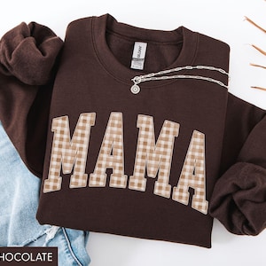 May include: Dark chocolate brown sweatshirt with the word "MAMA" in a plaid pattern across the front. A silver necklace with a pendant is draped across the sweatshirt. The text "DARK CHOCOLATE" is at the bottom.