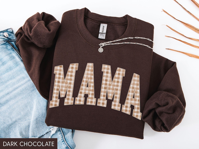May include: Dark chocolate brown sweatshirt with the word "MAMA" in a plaid pattern across the front. A silver necklace with a pendant is draped across the sweatshirt. The text "DARK CHOCOLATE" is at the bottom.