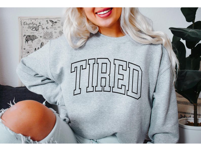 Pu&ograve; includere: Una felpa grigio chiaro con la scritta "TIRED" stampata a caratteri neri e in grassetto. La felpa ha un girocollo e maniche lunghe. La persona che indossa la felpa sta sorridendo.