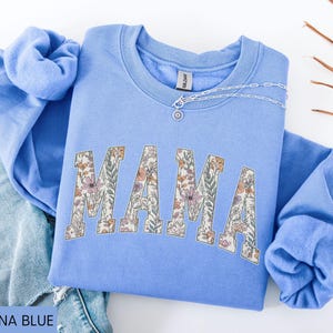 May include: Carolina blue sweatshirt with the word "MAMA" in floral print. A silver necklace with a pendant is on the neckline. The sweatshirt is folded on top of a pair of blue jeans.
