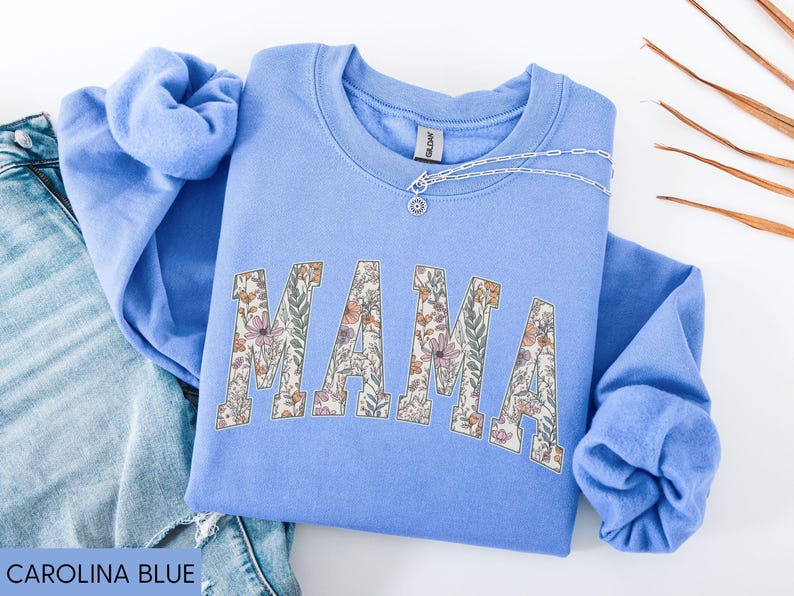 May include: Carolina blue sweatshirt with the word "MAMA" in floral print. A silver necklace with a pendant is on the neckline. The sweatshirt is folded on top of a pair of blue jeans.