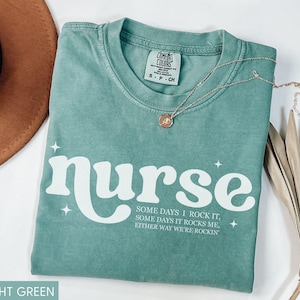 Pode incluir: Camiseta verde claro com a palavra "nurse" em branco, com uma fonte retr&ocirc;. A camiseta tamb&eacute;m apresenta o texto "Some days I rock it, some days it rocks me, either way we're rockin". Um colar de ouro e um chap&eacute;u marrom tamb&eacute;m est&atilde;o na imagem.
