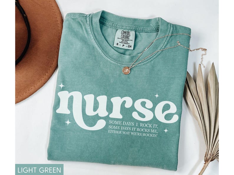 Pode incluir: Camiseta verde claro com a palavra "nurse" em branco, com uma fonte retr&ocirc;. A camiseta tamb&eacute;m apresenta o texto "Some days I rock it, some days it rocks me, either way we're rockin". Um colar de ouro e um chap&eacute;u marrom tamb&eacute;m est&atilde;o na imagem.