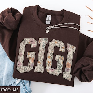 May include: A dark chocolate-coloured sweatshirt featuring the name "GIGI" in large, floral-patterned letters. A silver necklace with a pendant rests on the sweatshirt. The sweatshirt is folded next to a pair of blue jeans.