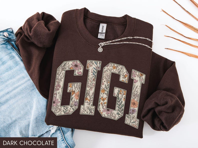 May include: A dark chocolate-coloured sweatshirt featuring the name "GIGI" in large, floral-patterned letters. A silver necklace with a pendant rests on the sweatshirt. The sweatshirt is folded next to a pair of blue jeans.