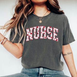 Pode incluir: Uma camiseta cinza escura com um gr&aacute;fico branco e rosa que diz "NURSE" em um padr&atilde;o de cora&ccedil;&atilde;o.