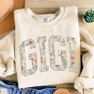 May include: A sand-coloured sweatshirt featuring the word "GIGI" in large letters filled with floral patterns. The flowers are in shades of pink, orange, and purple. The sweatshirt is folded on a wooden surface.