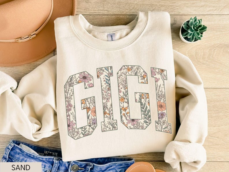 May include: A sand-coloured sweatshirt featuring the word "GIGI" in large letters filled with floral patterns. The flowers are in shades of pink, orange, and purple. The sweatshirt is folded on a wooden surface.