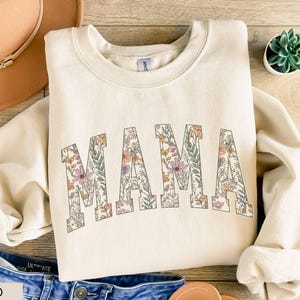 May include: A sand-colored sweatshirt with the word "MAMA" in large, floral-patterned letters. The floral design includes pink, orange, and green elements. The sweatshirt is paired with blue jeans and a tan hat.