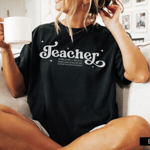 May include: Black t-shirt with the word "Teacher" in a white, stylized font. The shirt also features the text "Some days I rock it, some days it rocks me, either way we're rockin". The shirt is a casual style.