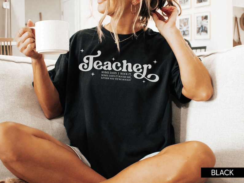 May include: Black t-shirt with the word "Teacher" in a white, stylized font. The shirt also features the text "Some days I rock it, some days it rocks me, either way we're rockin". The shirt is a casual style.