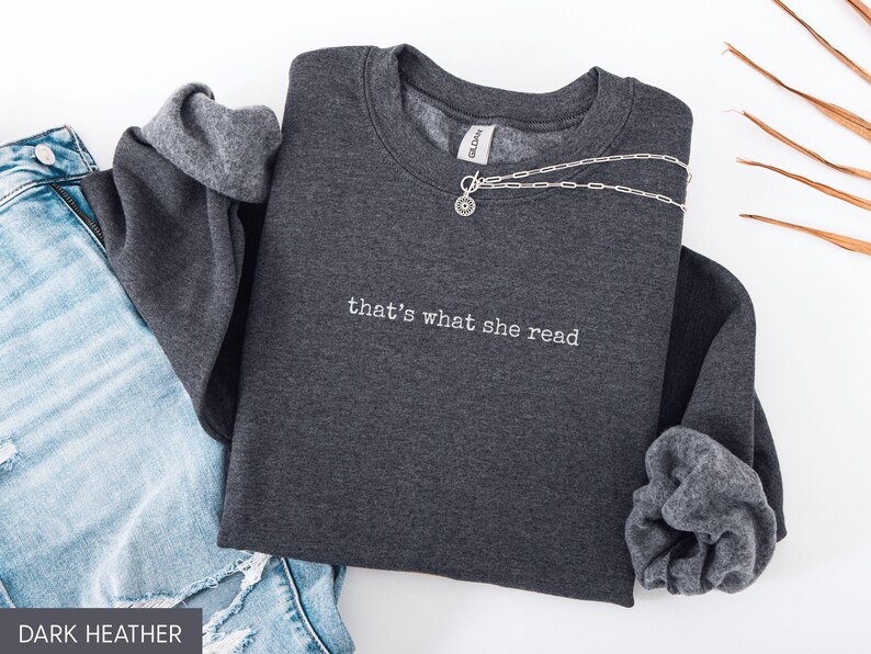 May include: Dark heather gray sweatshirt with the embroidered text "that's what she read." A silver necklace is draped across the front. The sweatshirt is paired with blue jeans. The Gildan brand tag is visible.