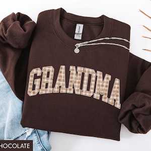 May include: Dark chocolate brown sweatshirt with the word "GRANDMA" in a plaid design. A silver necklace with a pendant is draped across the sweatshirt. The sweatshirt is paired with blue jeans.