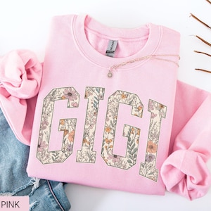 May include: A pale pink sweatshirt featuring the word "GIGI" in large, floral-filled letters. The letters are in a collegiate style, filled with a pattern of flowers and foliage in shades of pink, orange, and green. A delicate necklace rests on the sweatshirt.