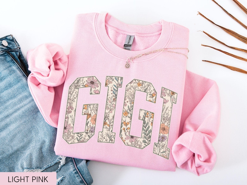 May include: A pale pink sweatshirt featuring the word "GIGI" in large, floral-filled letters. The letters are in a collegiate style, filled with a pattern of flowers and foliage in shades of pink, orange, and green. A delicate necklace rests on the sweatshirt.