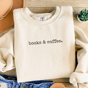 May include: A sand-colored sweatshirt with the text "books & coffee" in black. The sweatshirt is laid on a wooden surface with a hat, jeans, and a small potted plant. The sleeves are cuffed.