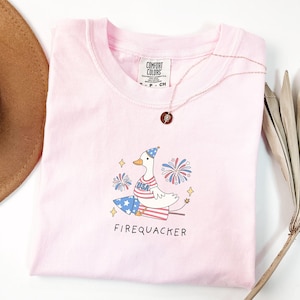 Op de afbeelding: Roze T-shirt met een cartoon eend die een rood, wit en blauw hoedje draagt en een vuurwerk vasthoudt. De tekst "Firequacker" staat onder de eend.