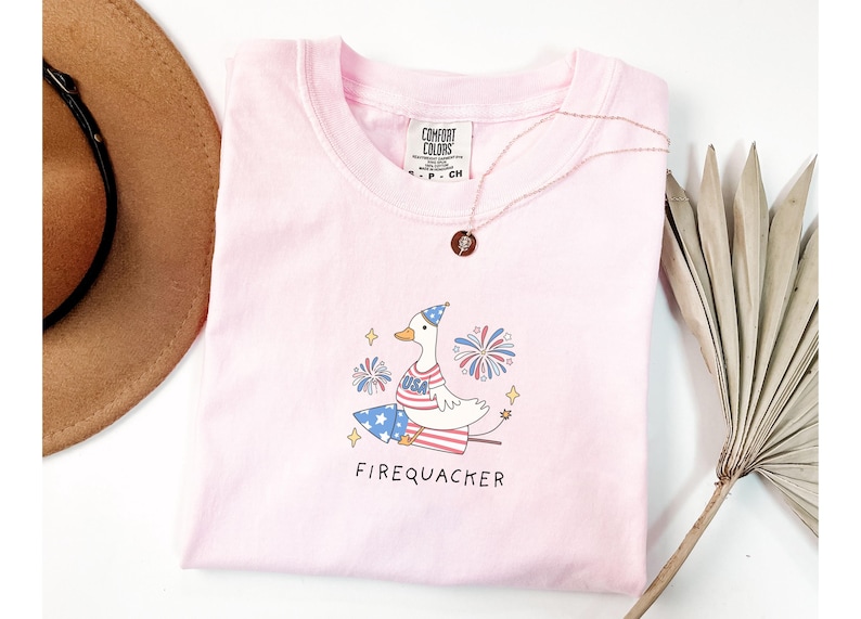 Op de afbeelding: Roze T-shirt met een cartoon eend die een rood, wit en blauw hoedje draagt en een vuurwerk vasthoudt. De tekst "Firequacker" staat onder de eend.
